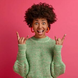 Happy carefree dark skinned rebellious young woman enjoys awesome music makes rock n roll gesture has fun on music festival or cool event wears casual jumper poses against pink wall mobile app development